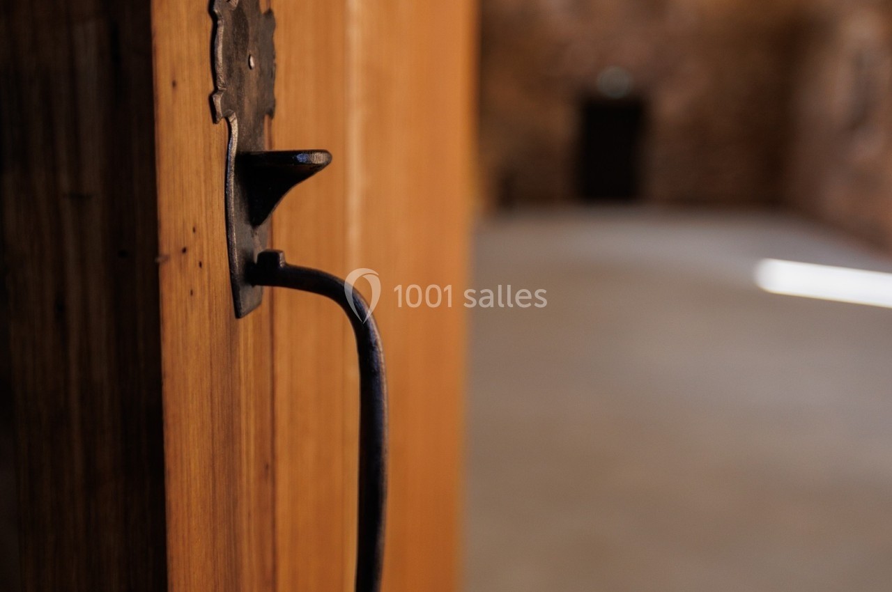 Porte en bois entrouverte avec une poignée en métal noir, donnant sur une pièce lumineuse aux murs en pierre.