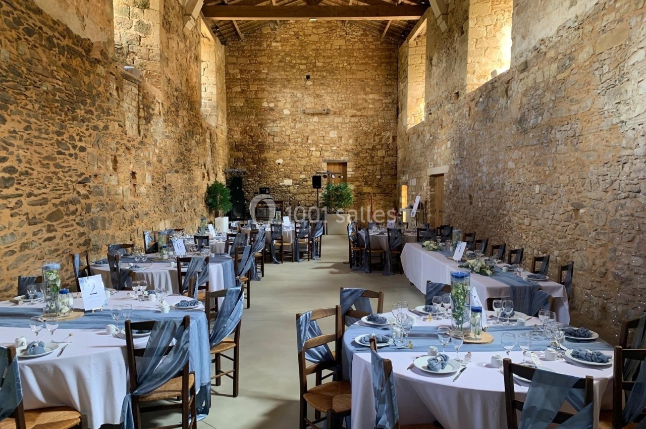 Salle en pierre avec tables rondes dressées pour un événement, décorées de nappes blanches et de chemins de table gris.