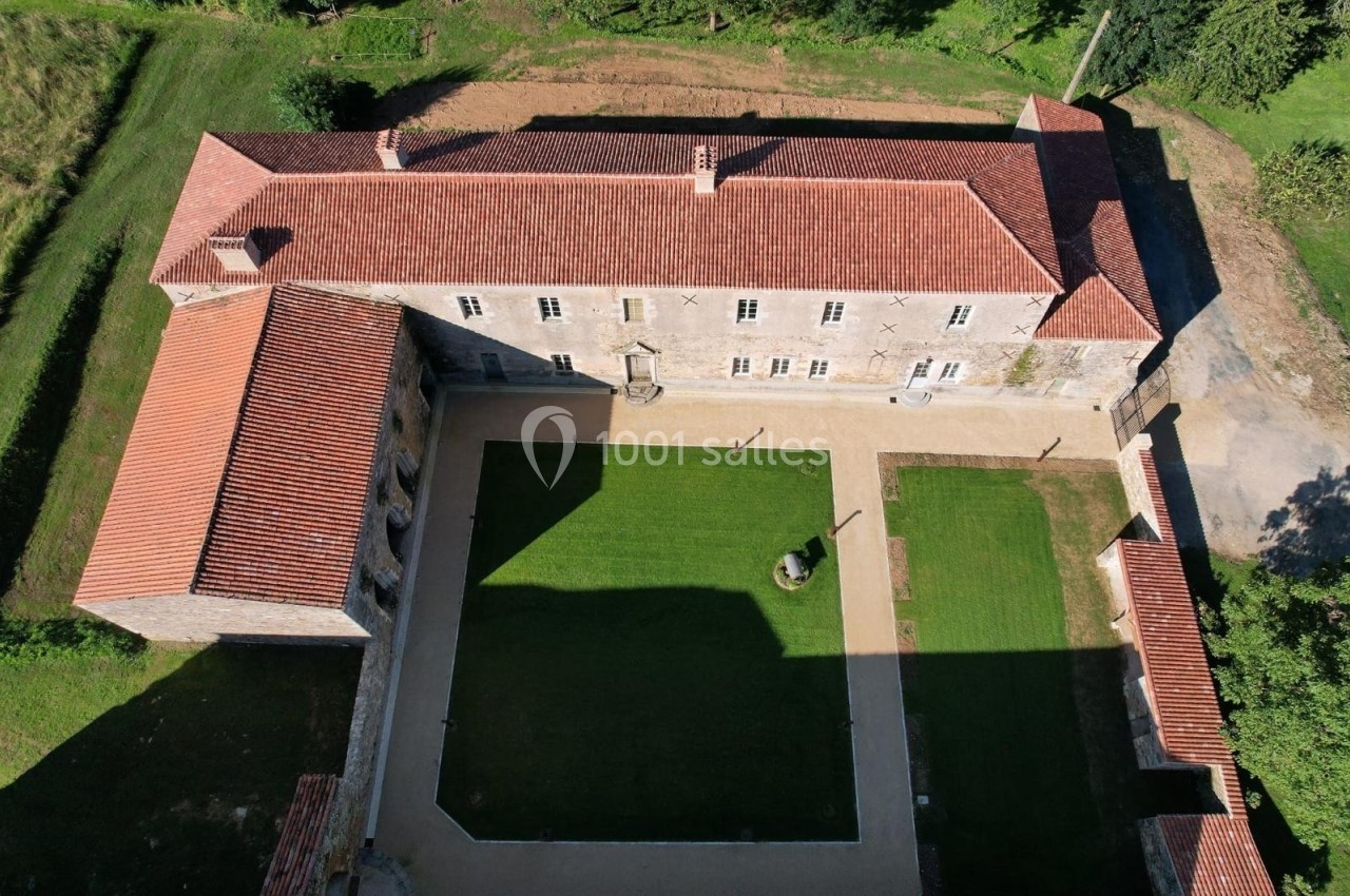 Vue aérienne d'un bâtiment ancien en pierre avec toit en tuiles rouges entourant une cour intérieure gazonnée.
