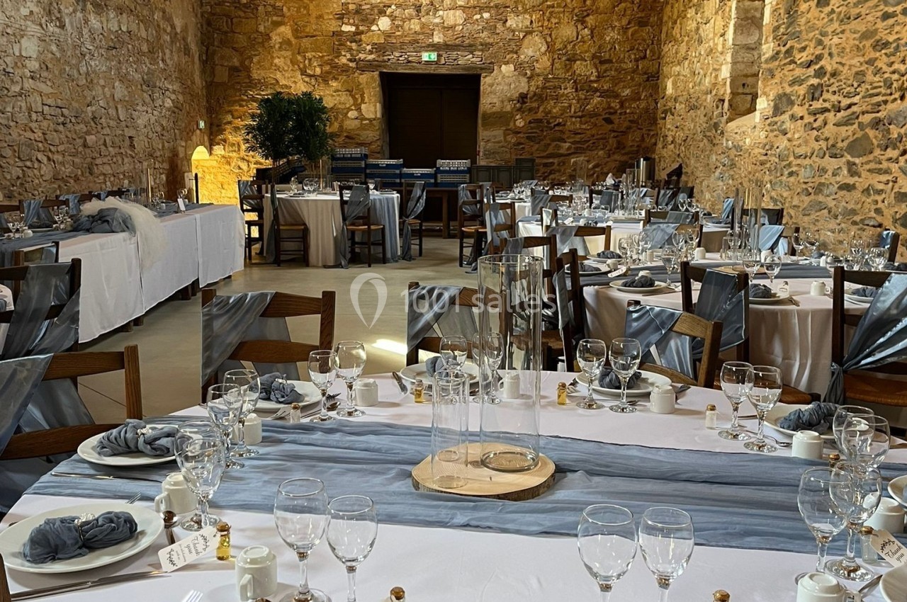 Salle en pierre ancienne aménagée pour un repas, avec tables dressées, nappes blanches et serviettes grises.