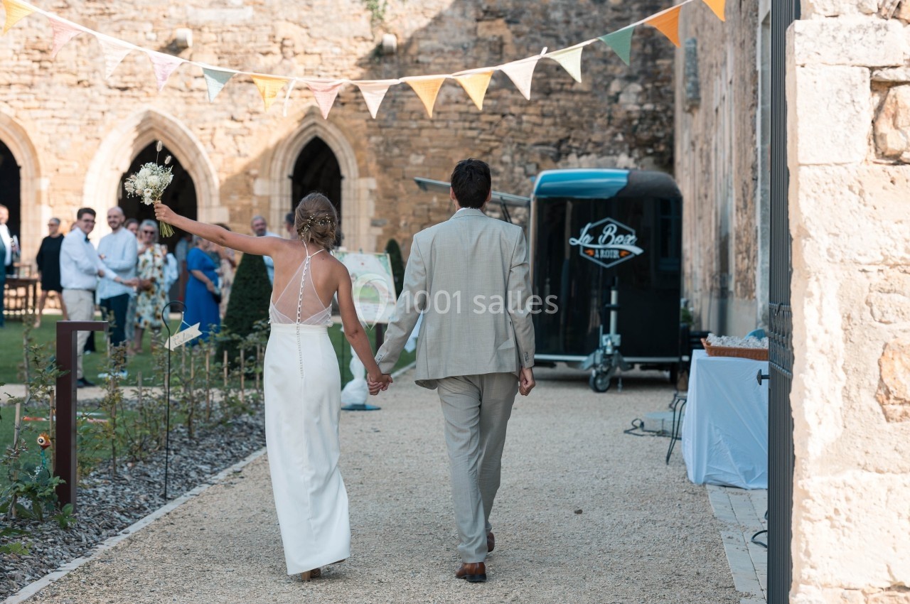 Un couple en tenue de mariage marche main dans la main dans une cour en pierre, décorée de fanions colorés.