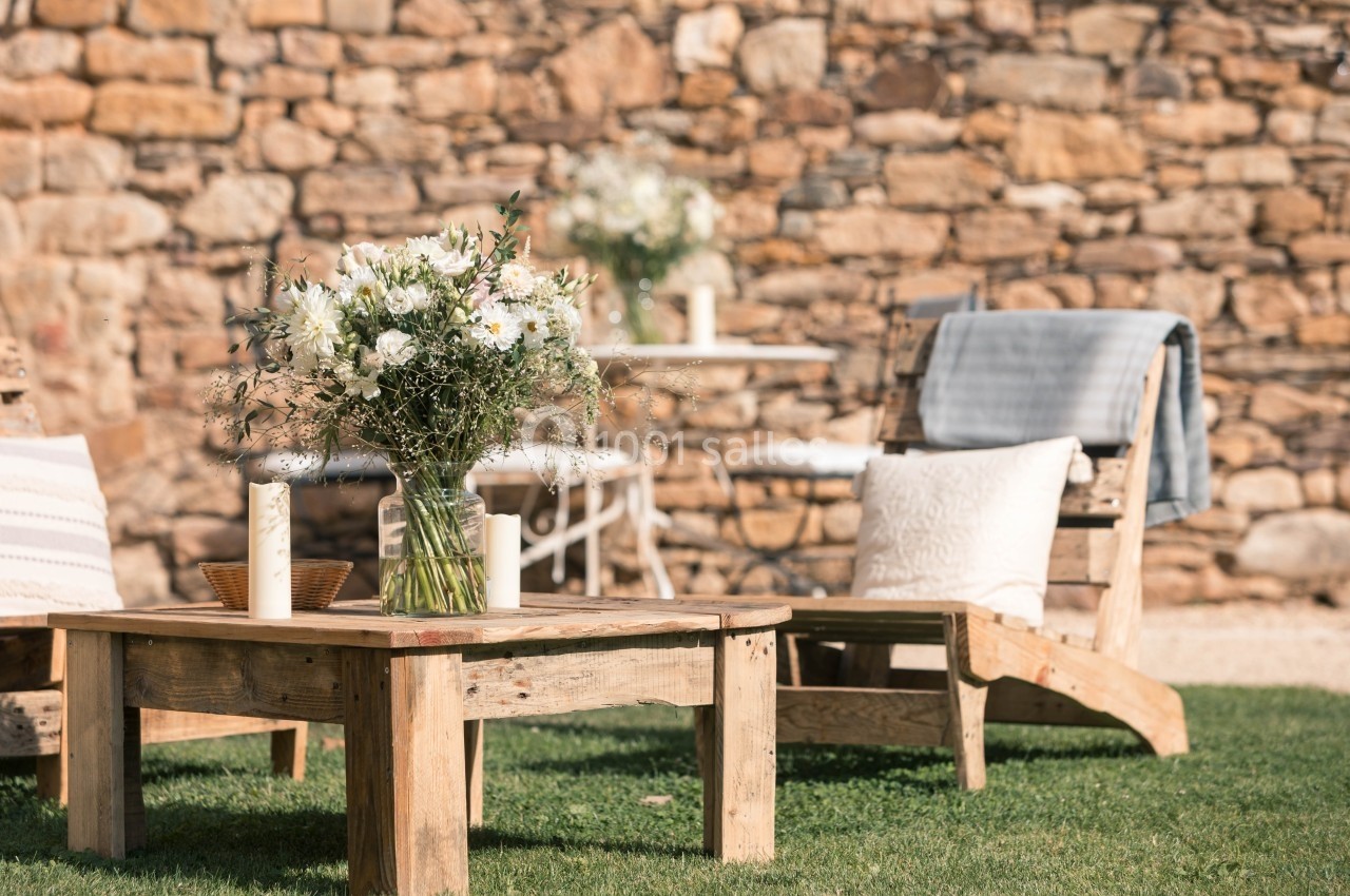 Table basse en bois avec un bouquet de fleurs blanches, entourée de chaises longues et d'une table sur pelouse devant un mur…