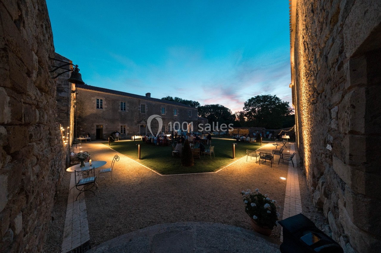 Cour intérieure d'un bâtiment en pierre éclairée à la tombée de la nuit, avec des tables et des invités rassemblés.
