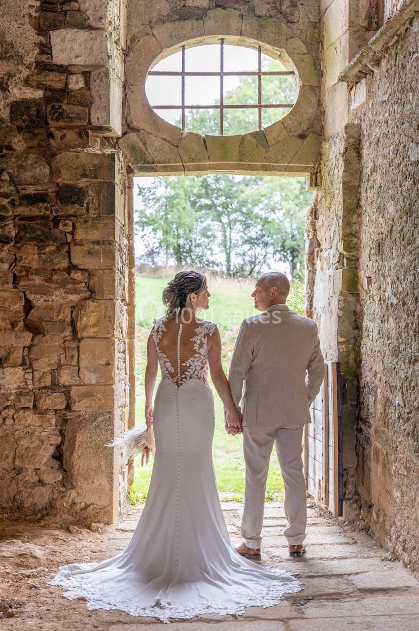 Un couple en tenue de mariage se tient la main dans une vieille bâtisse en pierre, avec vue sur un paysage verdoyant.