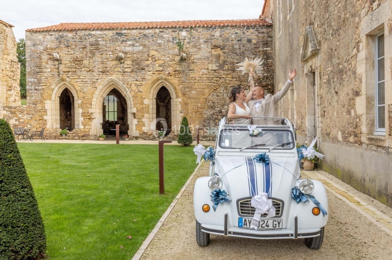 Un couple de mariés souriants dans une voiture ancienne décorée, devant un bâtiment en pierre historique.