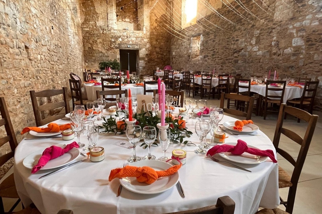 Salle en pierre décorée pour un repas festif, avec tables rondes dressées, serviettes colorées et bougies roses.