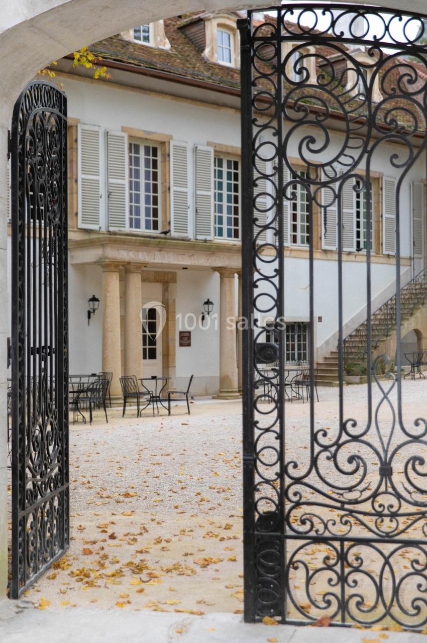 Portail en fer forgé ouvert donnant sur une cour pavée avec des tables, des chaises et une maison aux volets blancs.