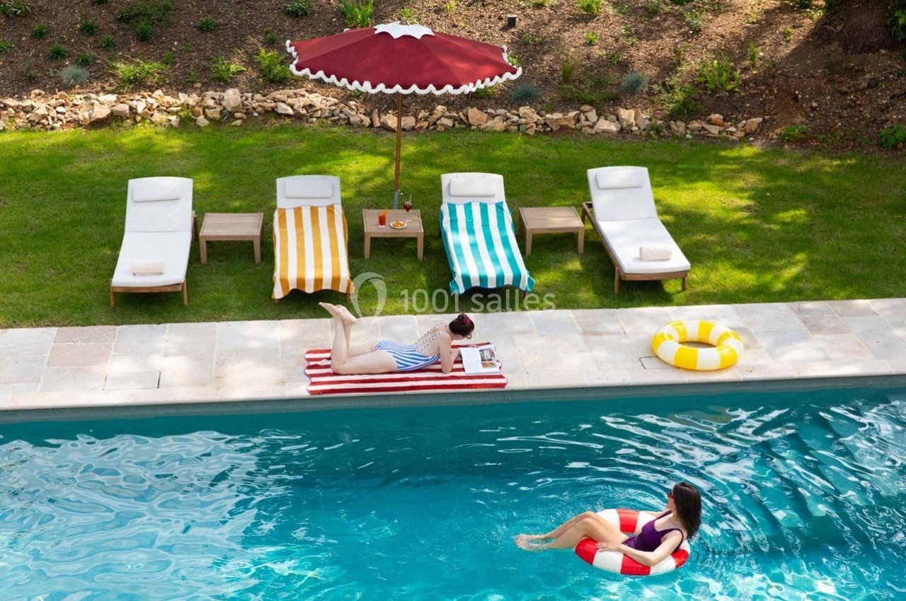 Deux personnes profitent d'une piscine, l'une nageant avec une bouée, l'autre allongée sur une chaise longue près de l'eau.