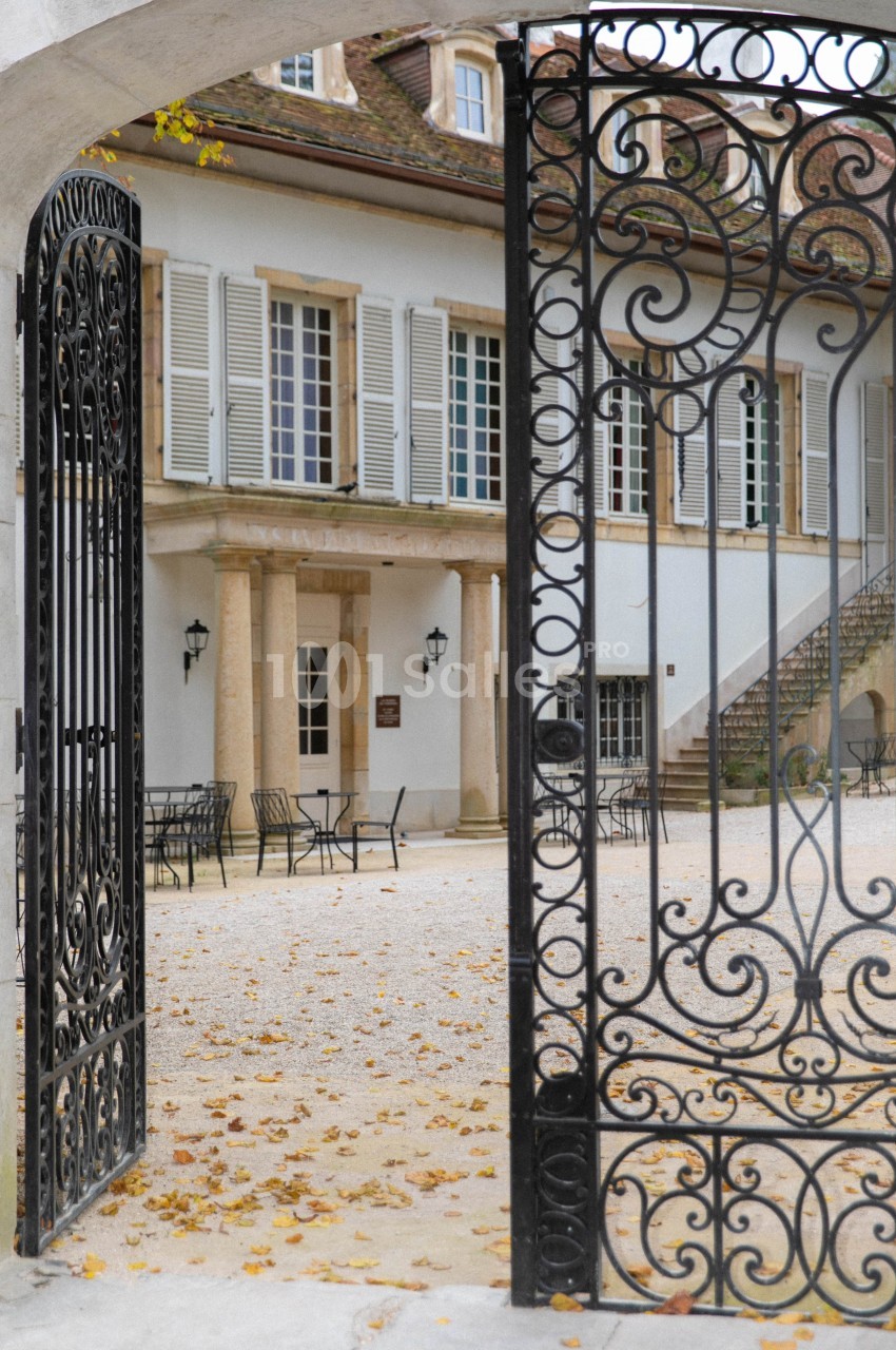 Portail en fer forgé ouvert donnant sur une cour pavée avec des tables, des chaises et une bâtisse aux volets blancs.