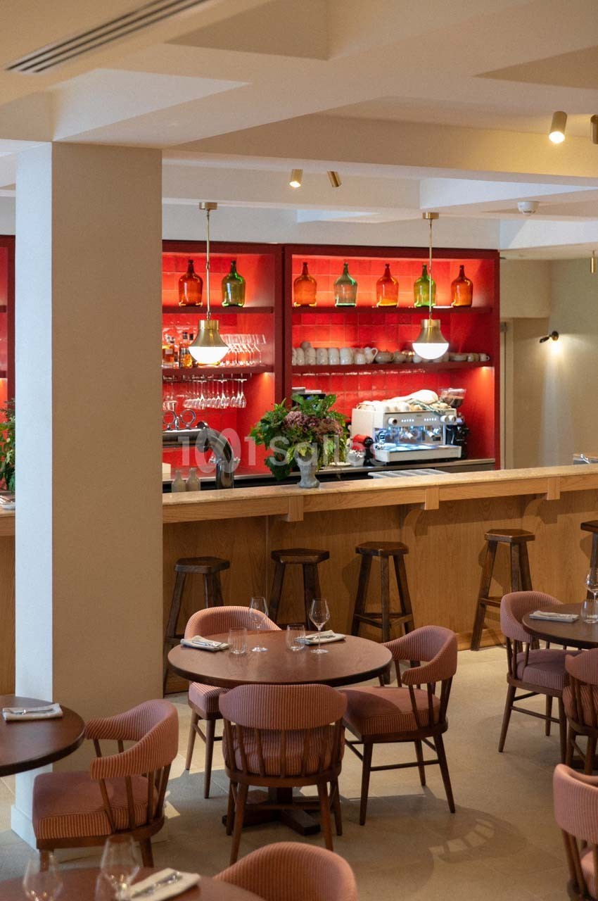 Salle de restaurant avec tables dressées, chaises en bois et un bar en arrière-plan avec étagères rouges et éclairage…