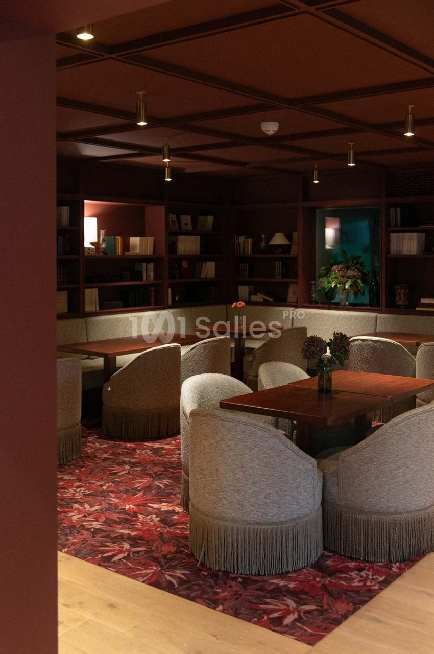 Salle de lecture chaleureuse avec fauteuils en tissu, tables en bois, étagères remplies de livres et éclairage tamisé.