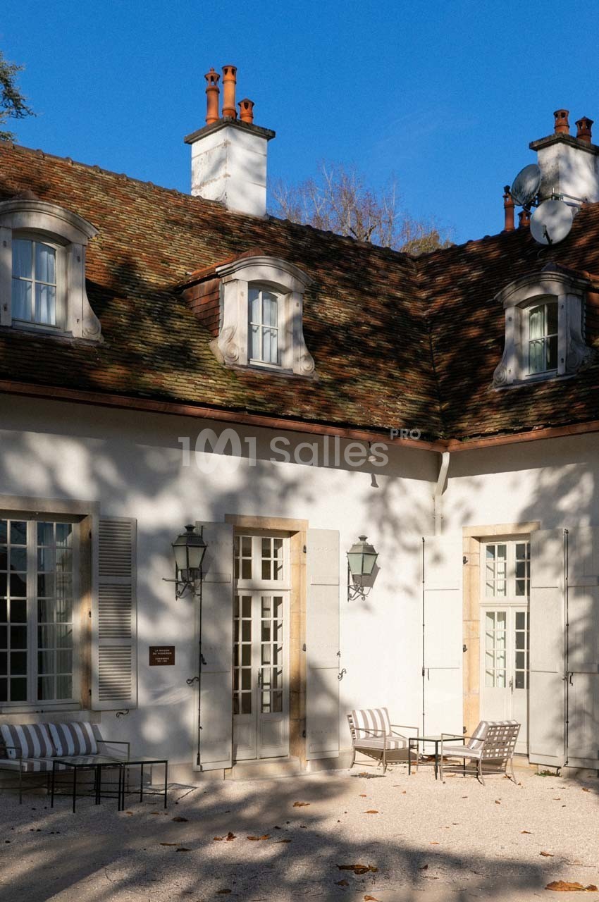 Façade d'une maison avec toit en tuiles, fenêtres mansardées et terrasse équipée de chaises sous un ciel bleu.