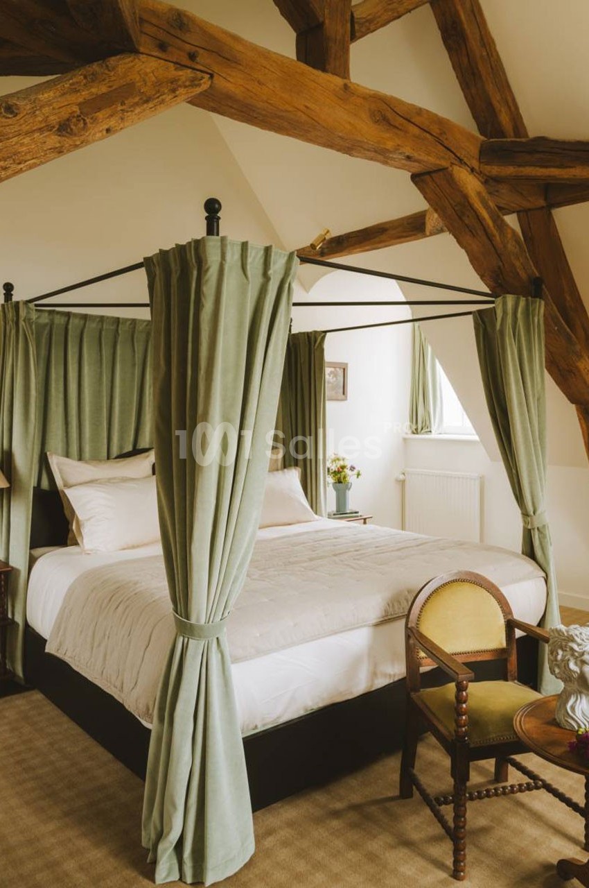 Lit à baldaquin avec rideaux verts dans une chambre lumineuse aux poutres apparentes et mobilier classique.