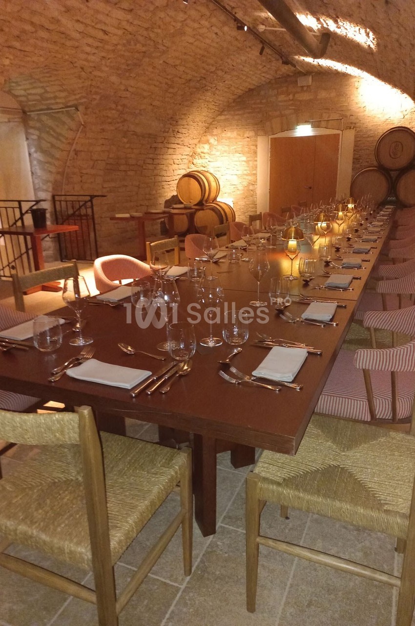 Grande table en bois dressée avec vaisselle et verres, située dans une cave voûtée avec des tonneaux en arrière-plan.
