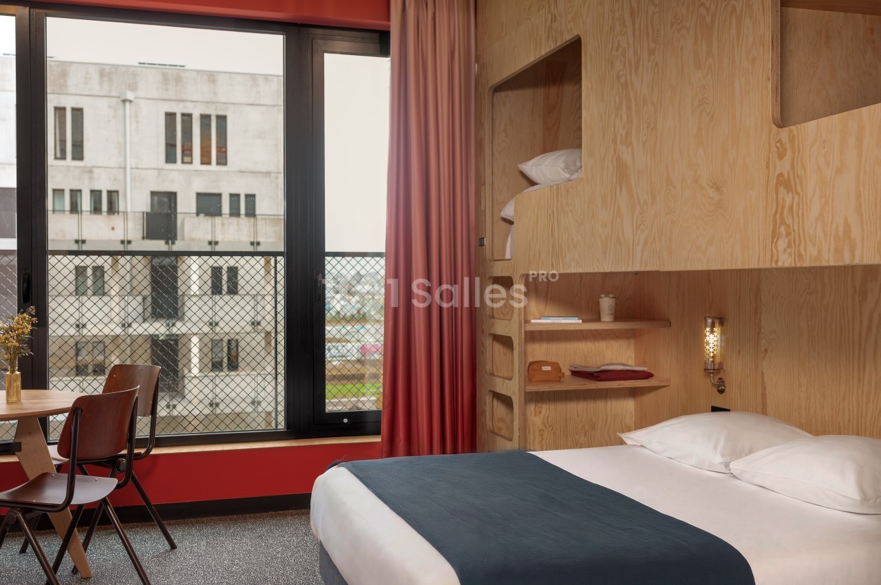 Chambre d'hôtel moderne avec lit double, étagères en bois, table et chaises, vue sur un bâtiment extérieur.