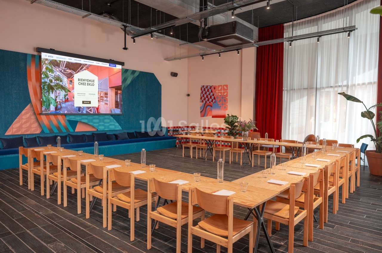 Salle de réunion lumineuse avec tables en U, chaises en bois, écran de projection et décoration colorée.
