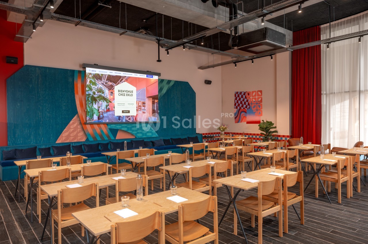 Salle de conférence avec des rangées de tables et chaises en bois, écran de projection allumé et décoration colorée.