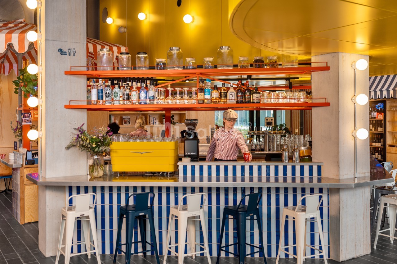 Bar coloré avec comptoir rayé bleu et blanc, étagères orange, décorations lumineuses et une personne servant une boisson.