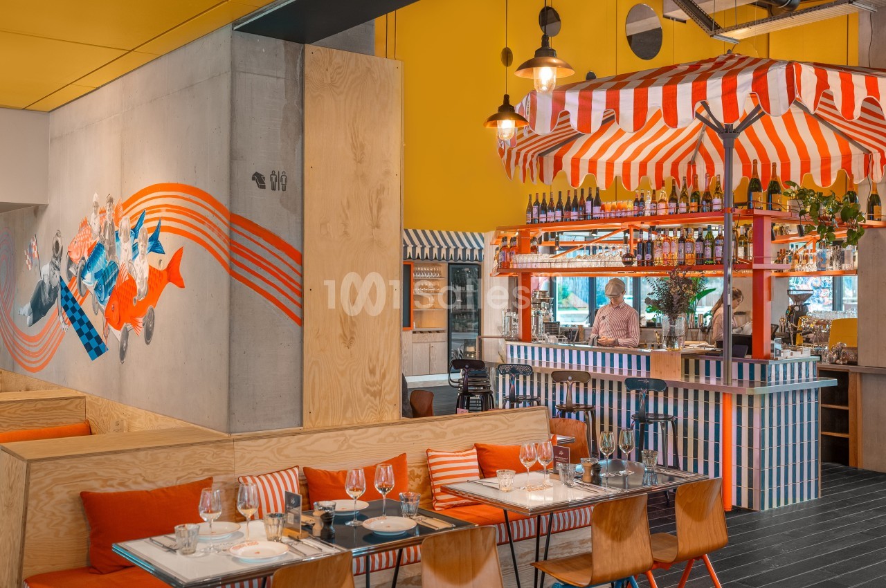 Intérieur d'un restaurant coloré avec un bar à rayures rouges et blanches, des tables dressées et une fresque murale.