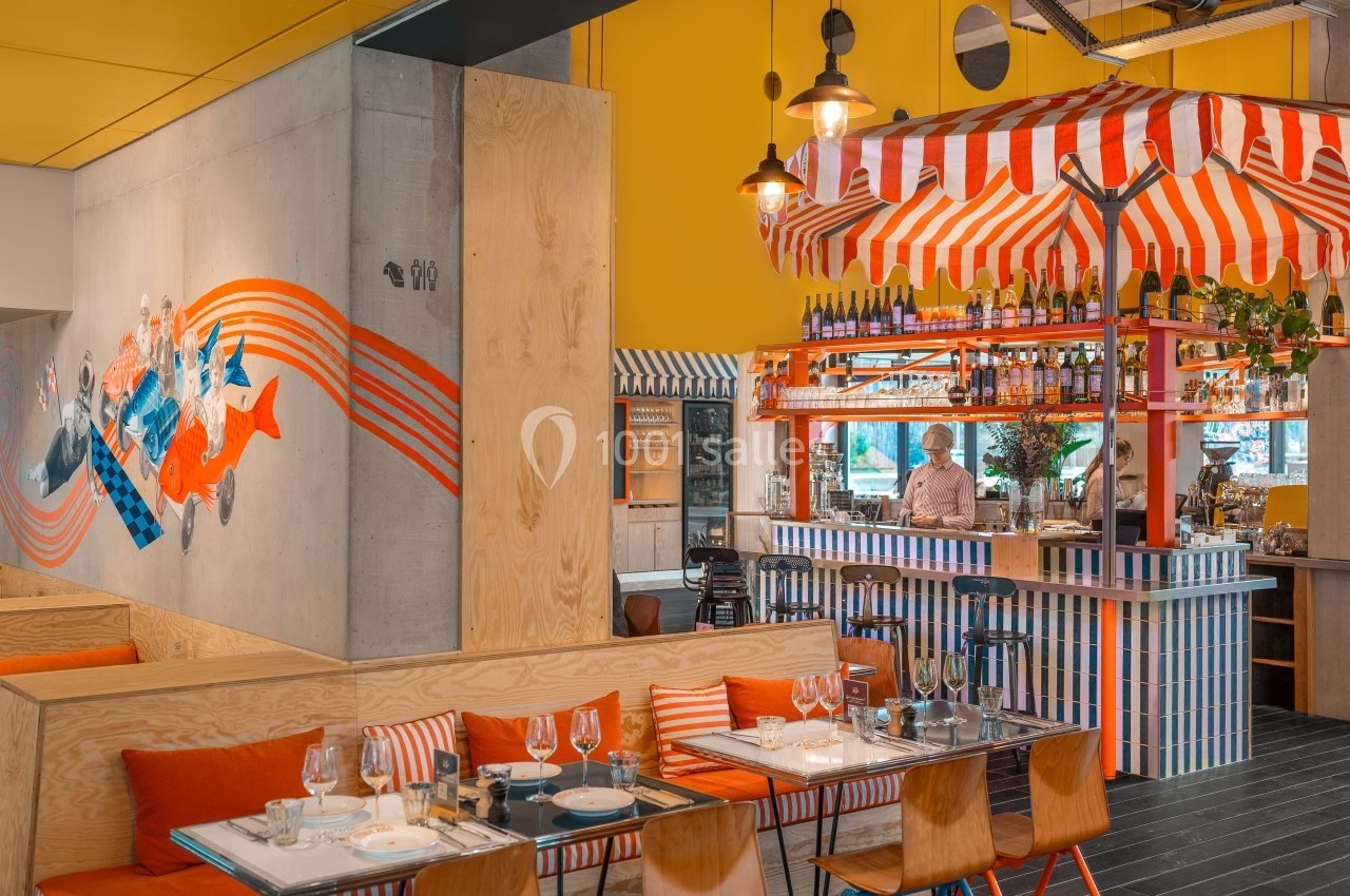Intérieur d'un restaurant coloré avec un bar à rayures rouges et blanches, des tables dressées et une fresque murale.