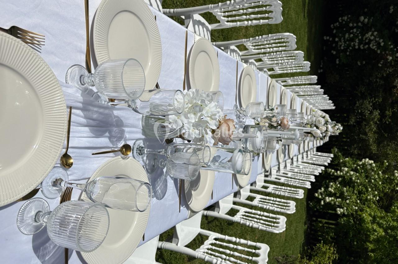 Table dressée en extérieur avec nappes blanches, assiettes, verres, couverts dorés et bouquets de fleurs au centre.