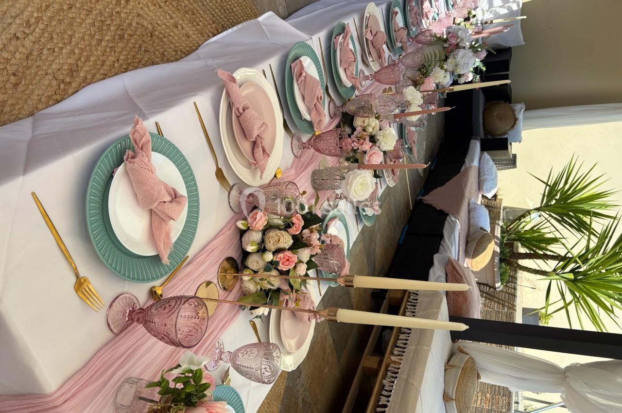 Table dressée avec nappes blanches, vaisselle pastel, décorations florales roses et bougies dans un cadre lumineux.