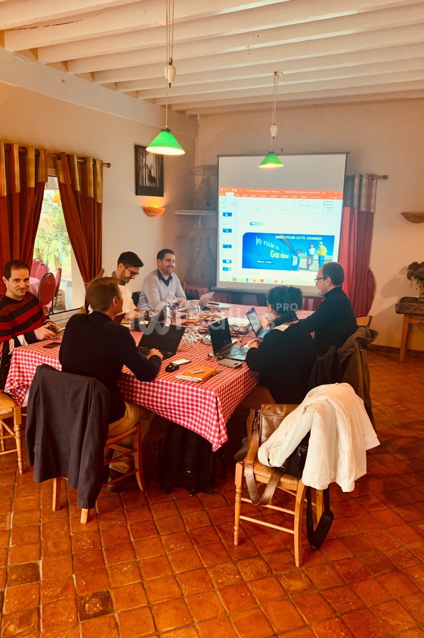 Des personnes travaillent autour d'une table avec ordinateurs, dans une salle équipée d'un projecteur et d'une décoration…