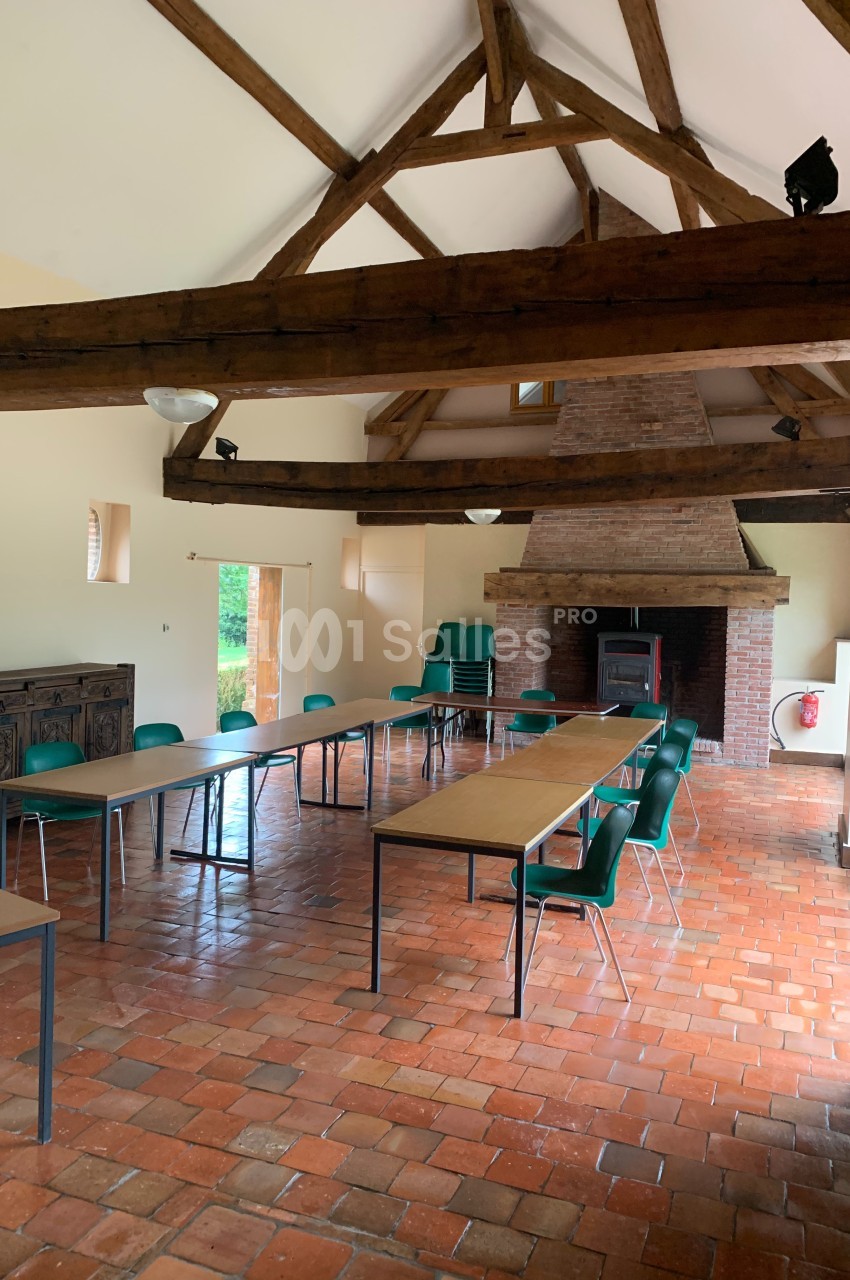 Salle lumineuse avec poutres apparentes, tables alignées, chaises vertes et cheminée en briques au fond.