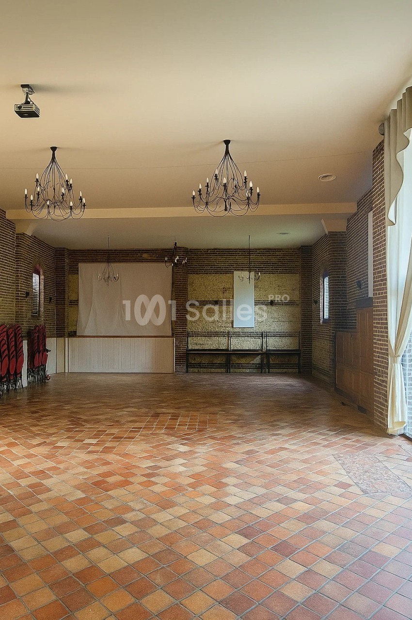 Salle vide avec sol en carreaux de terre cuite, murs en briques, rideaux ouverts et lustres suspendus.