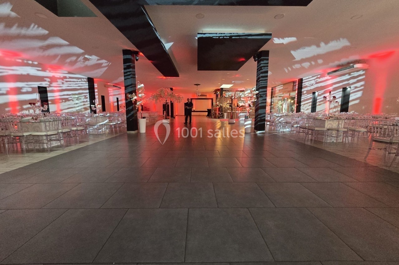 Salle de réception spacieuse avec éclairage rouge et blanc, tables décorées et chaises transparentes alignées.