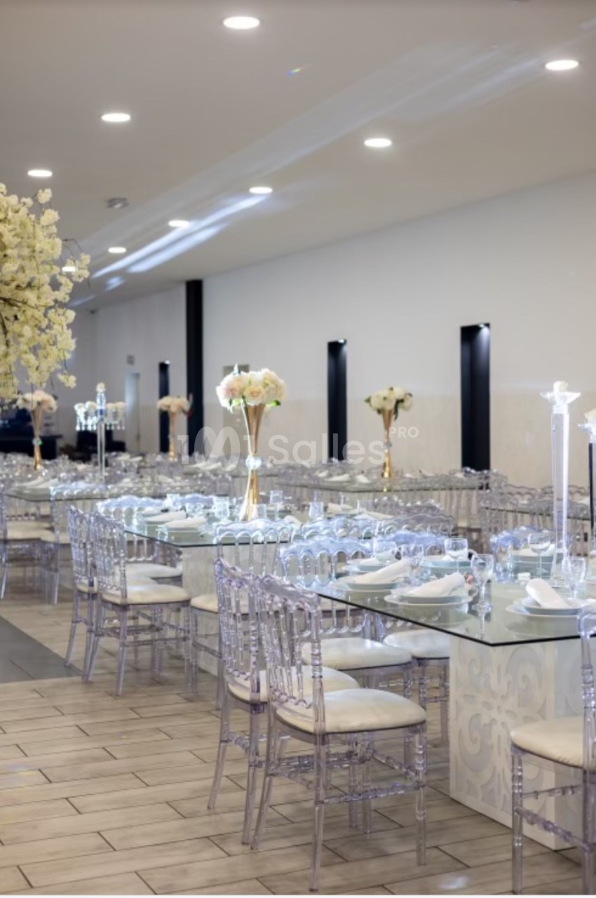 Salle de réception élégante avec tables dressées, chaises transparentes et décorations florales blanches.