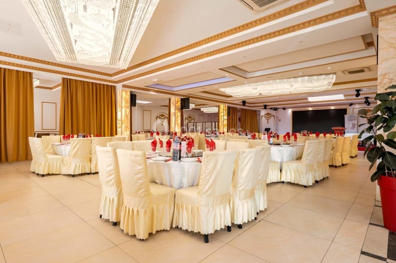 Salle de réception élégante avec tables rondes dressées, nappes blanches et chaises couvertes, éclairée par des lustres.