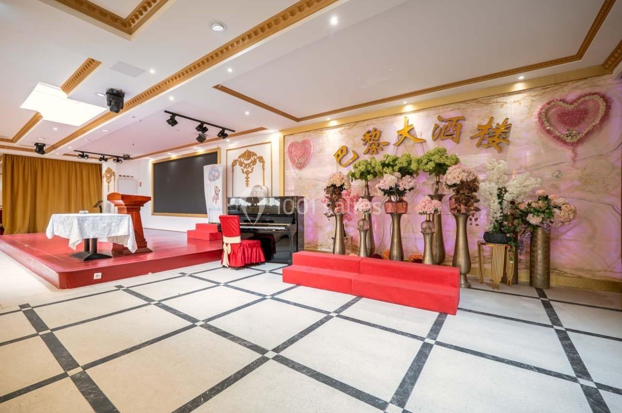 Salle décorée avec des fleurs, un piano noir, une scène avec pupitre et fond orné de caractères chinois dorés.