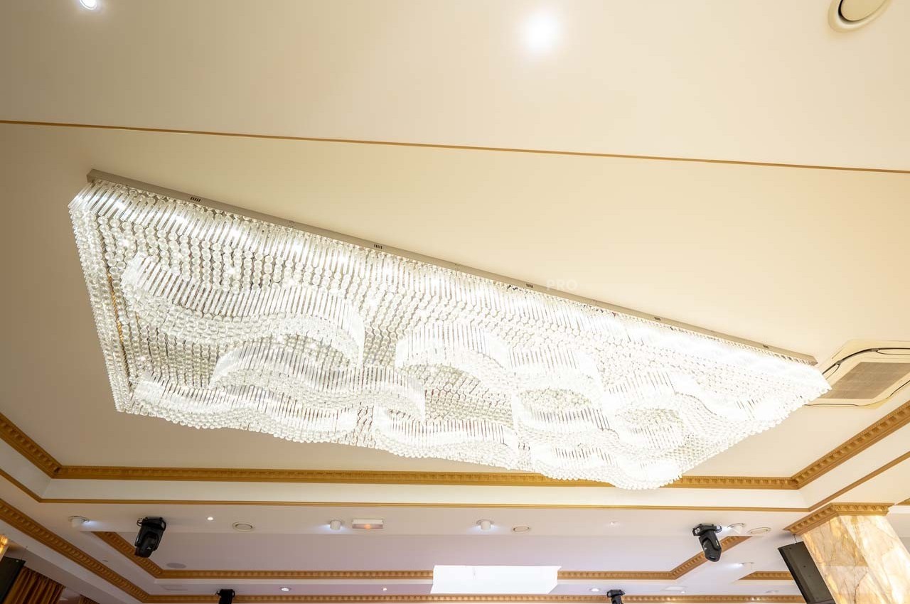 Plafonnier rectangulaire en cristal suspendu, éclairé, avec un design ondulé, dans une salle au plafond beige.