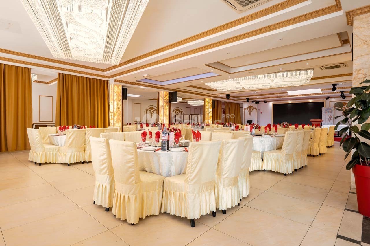 Salle de réception élégante avec tables rondes dressées, nappes blanches et chaises couvertes, éclairée par des lustres.
