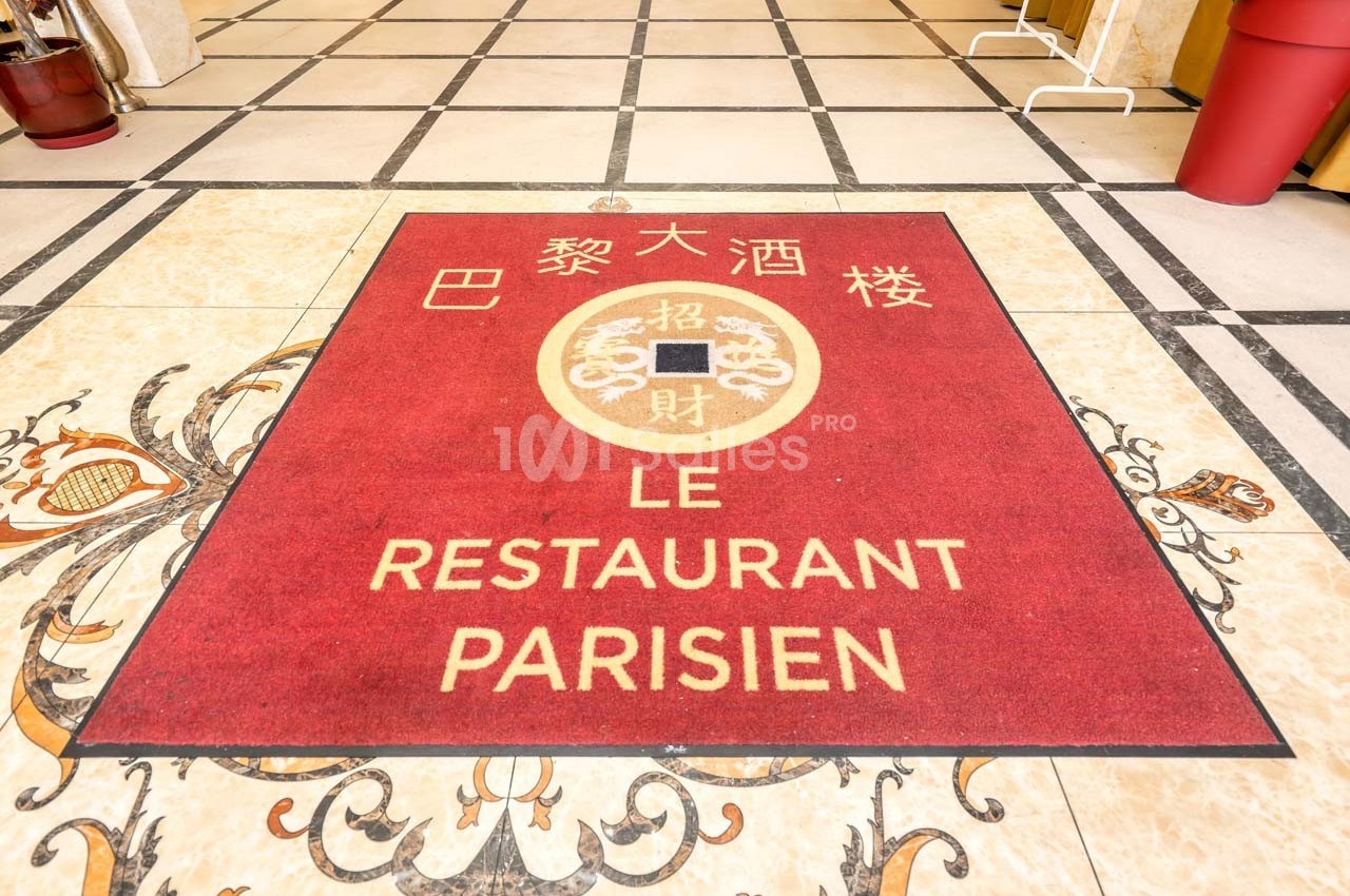 Tapis rouge avec inscriptions en français et en chinois, placé sur un sol carrelé à l'entrée d'un établissement.