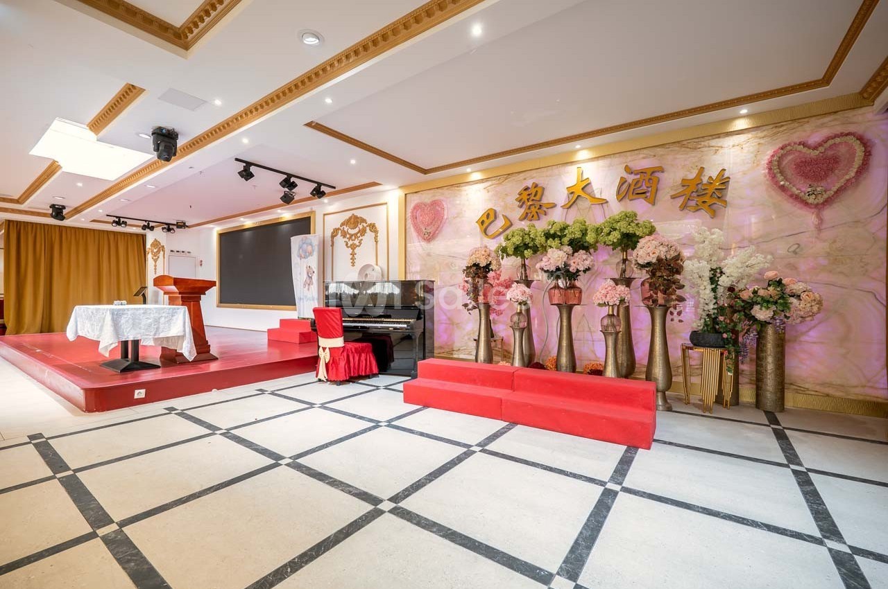Salle décorée avec des fleurs, un piano noir, une scène avec pupitre et fond orné de caractères chinois dorés.