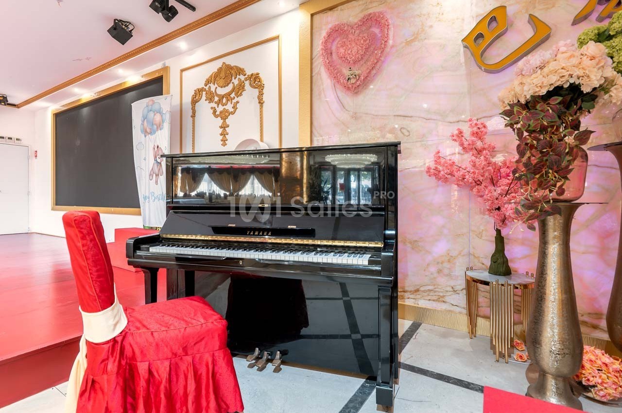 Piano droit noir devant une scène, entouré de décorations florales et d'éléments dorés sur un fond coloré.