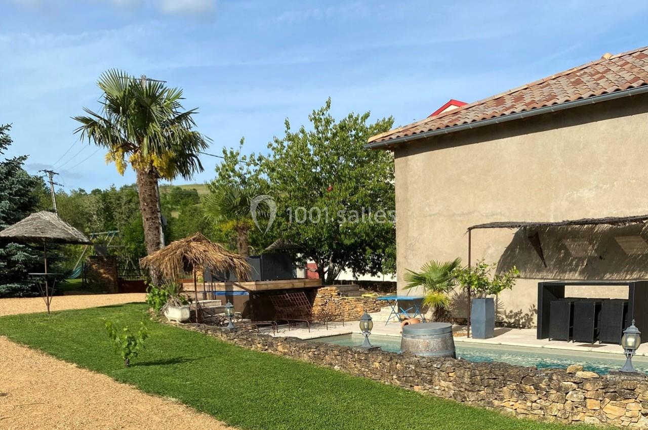 Jardin avec piscine, terrasse en bois, parasols en chaume, palmiers et mur en pierre sous un ciel dégagé.