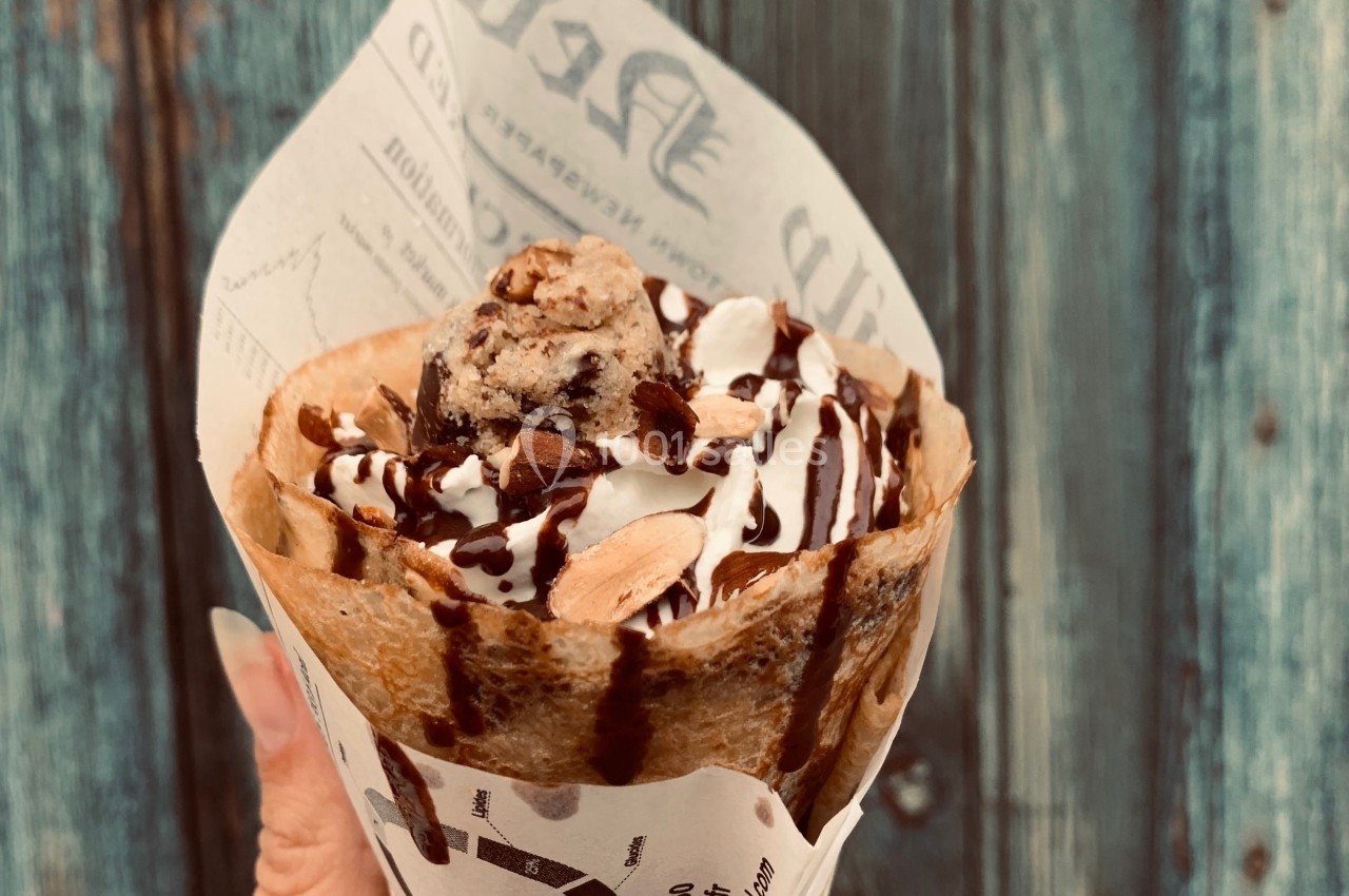 Crêpe garnie de crème, morceaux de biscuit et filet de chocolat, tenue à la main devant une porte en bois bleu.