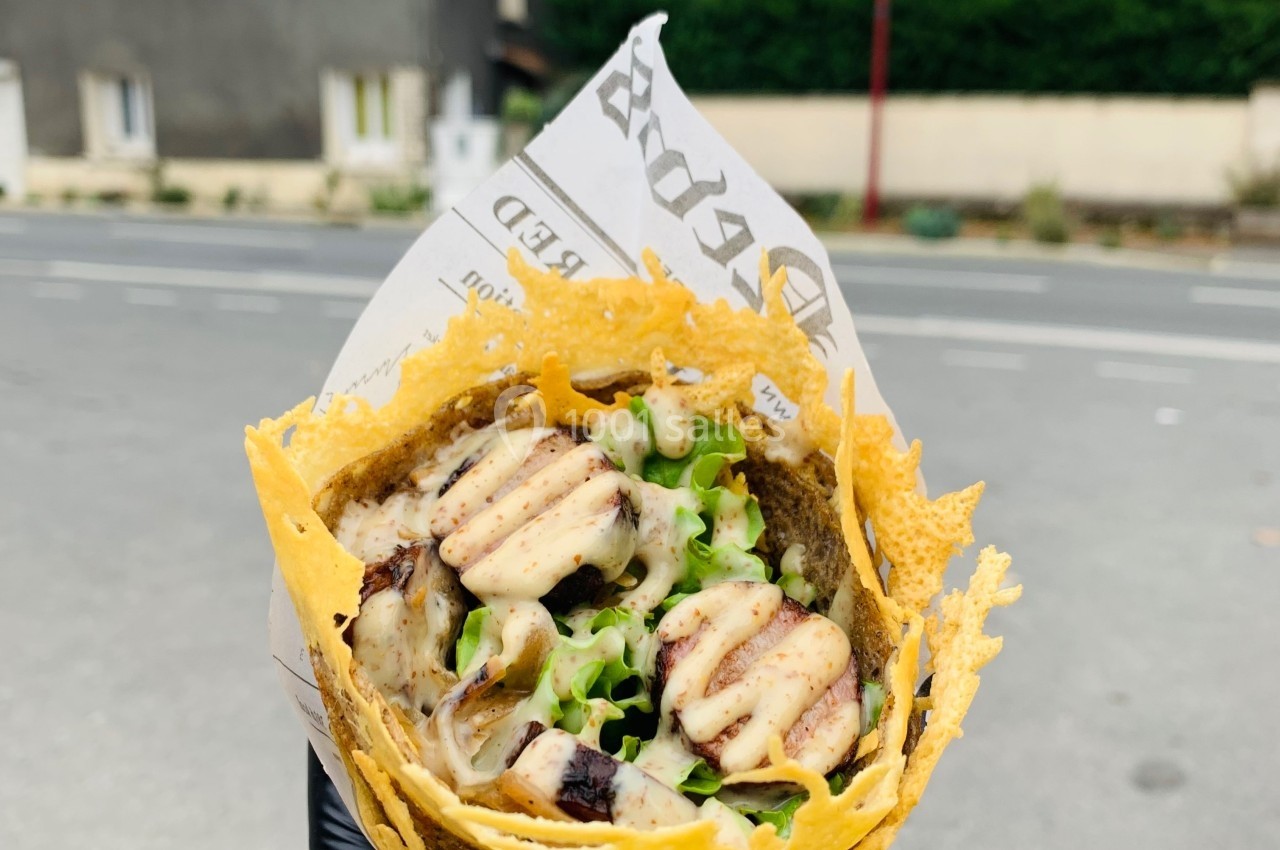 Galette garnie de légumes, viande grillée et sauce, tenue à la main avec un gant noir, en extérieur près d'une route.