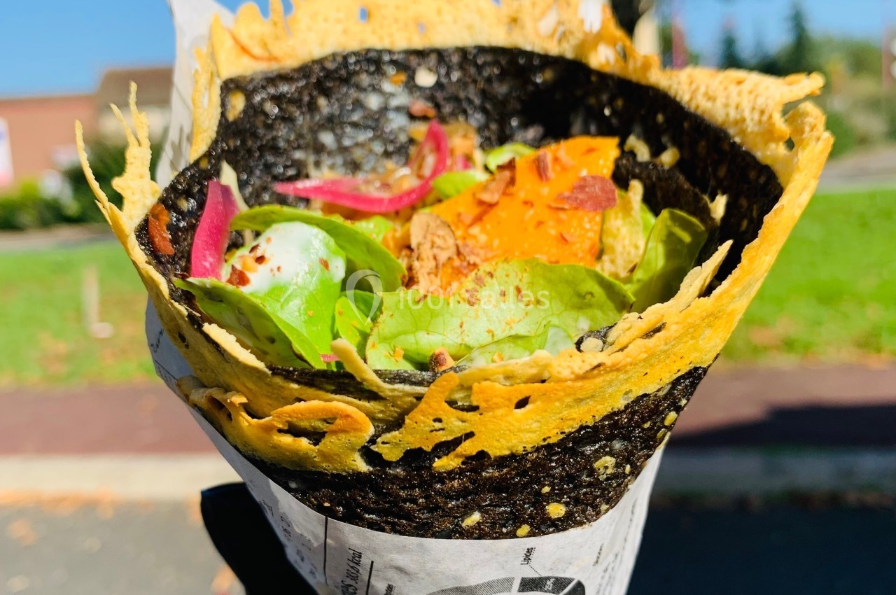 Tacos enroulé dans une galette noire et dorée, garni de légumes colorés, tenu par une main gantée en extérieur.