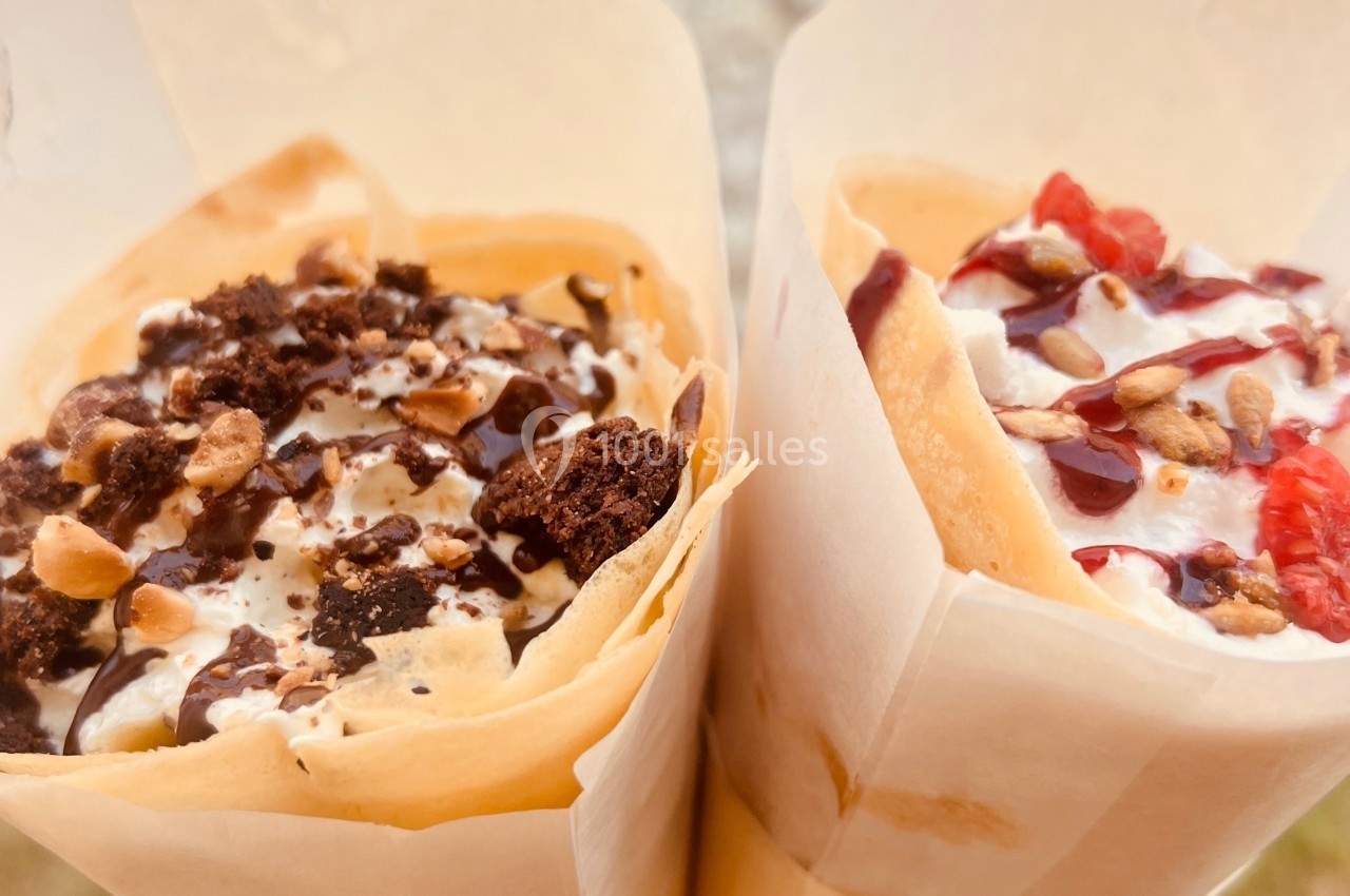 Deux crêpes garnies de crème, chocolat, éclats de noisettes et fruits rouges, tenues à la main en extérieur.