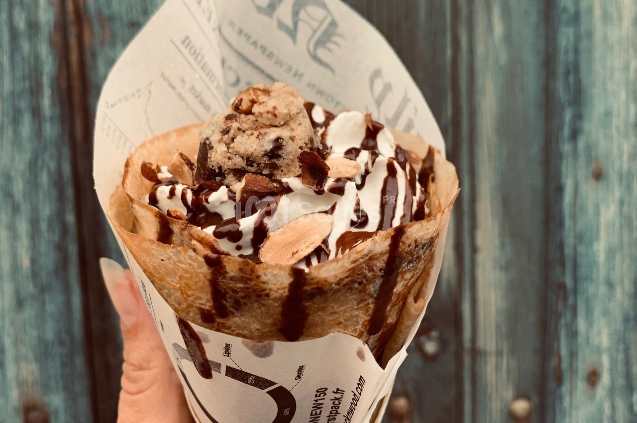 Crêpe garnie de glace, chantilly, amandes et sauce chocolat, présentée dans un papier imprimé devant un fond en bois.