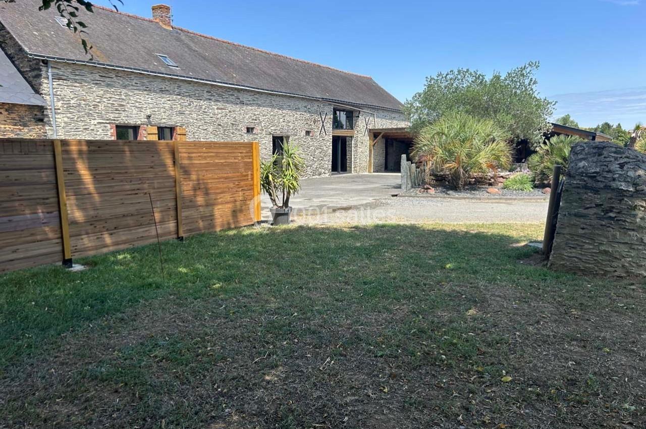 Cour devant une maison en pierre avec clôture en bois, plantes en pot et jardin arboré sous un ciel dégagé.
