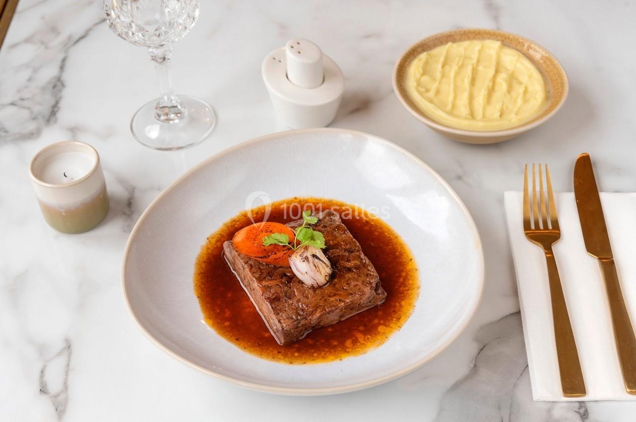 Plat de viande nappé de sauce, garni de légumes, accompagné de purée dans un bol sur une table dressée élégamment.