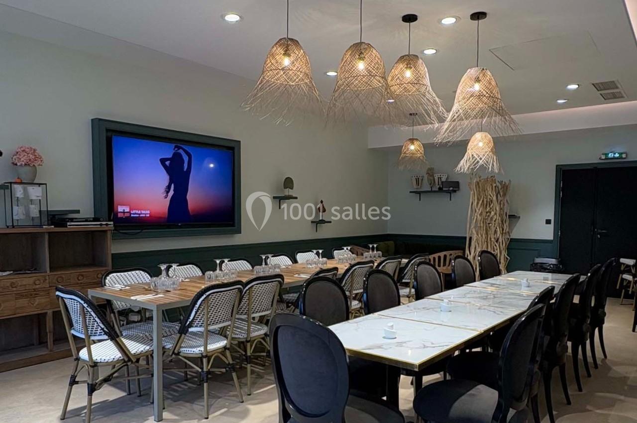Salle à manger lumineuse avec tables dressées, chaises variées et suspensions en osier, écran mural allumé.