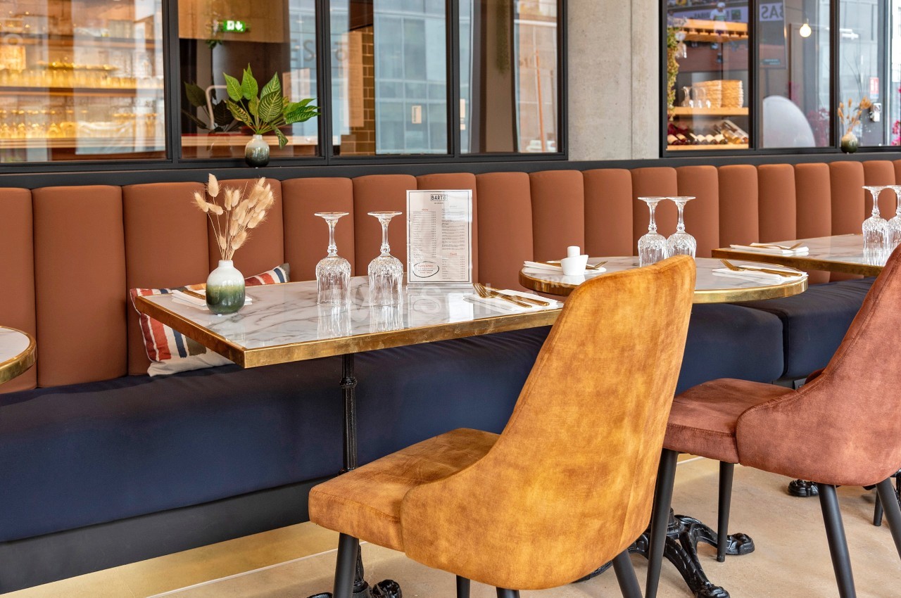 Salle de restaurant moderne avec tables en marbre, chaises rembourrées et banquette orange, décorée de plantes et de verres.