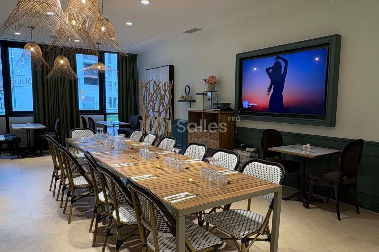 Salle de restaurant lumineuse avec une grande table en bois, des chaises en rotin et un écran affichant une silhouette au…