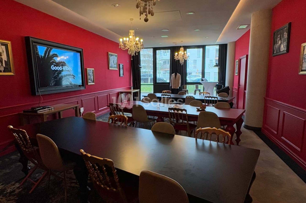 Salle de réunion lumineuse avec murs rouges, tables noires et chaises, équipée d'un écran et décorée de cadres.